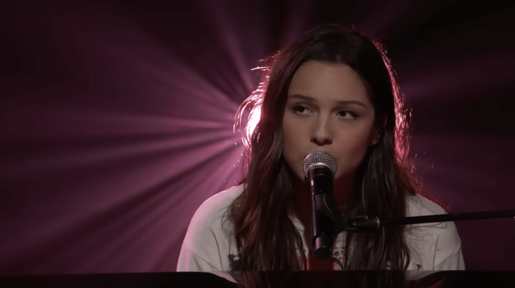 Olivia Rodrigo sitting, playing the piano while a spotlight shines on her from behind.