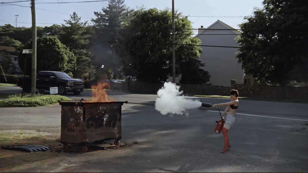 Gracie Abrams spraying a fire extinguisher at a burning dumpster on a suburban street, in a still from the 'Hit the Wall' music video.