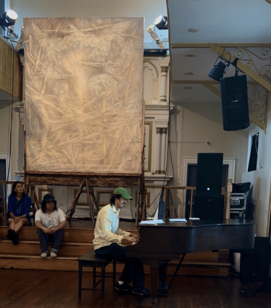 People sit on stairs as they watch Austin Kim performing at the piano. This scene is dwarfed by the massive painting in the background.