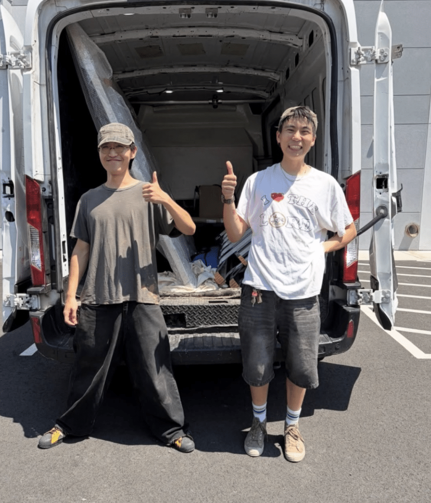 Both curators stand in front of an open van, with many different pieces of art and power tools in the background.