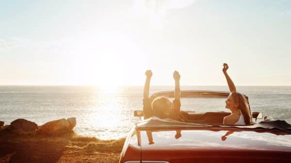 Two women in a convertible car at a beach on the coast of the ocean watching the sunset.