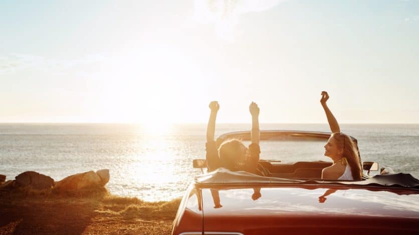 Two women in a convertible car at a beach on the coast of the ocean watching the sunset.